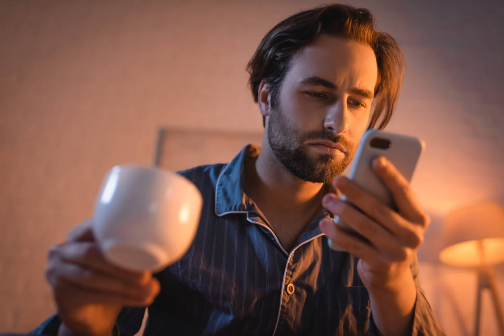 Молодий чоловік у піжамі тримає розмиту каву і смартфон вночі
  - Фото, зображення
