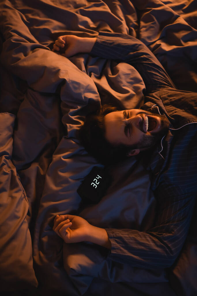 Вид сверху на человека в пижаме, зевающего возле будильника на кровати по ночам  - Фото, изображение