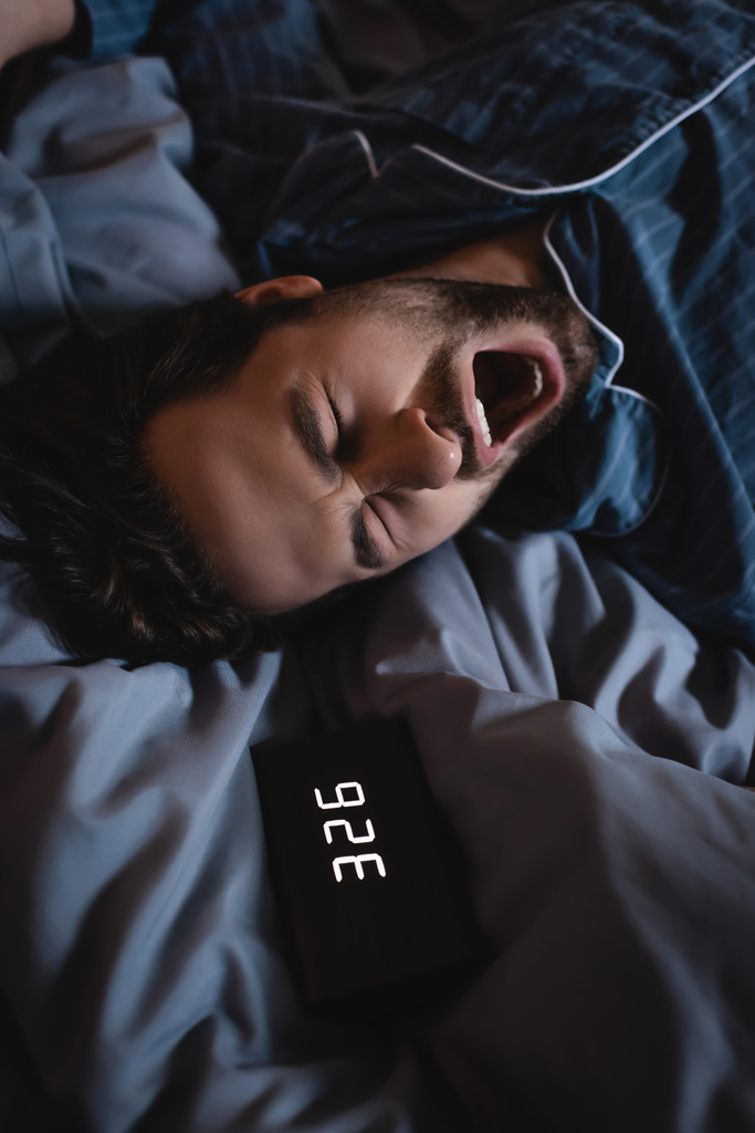 Vue de dessus de l'homme insomniaque bâillant près du réveil sur le lit la nuit  - Photo, image