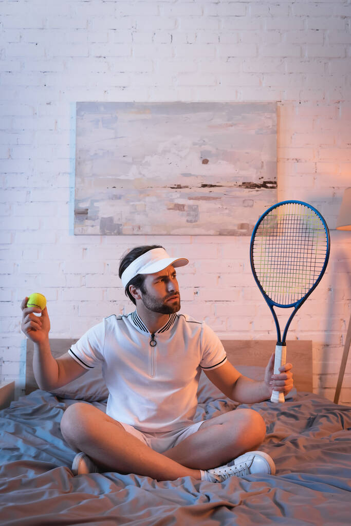Запутавшийся лунатик в спортивной одежде, держащий теннисный мяч и ракету на кровати  - Фото, изображение