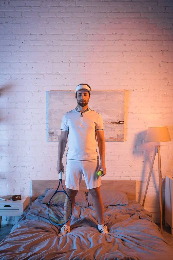 Sleepwalker in sportswear holding tennis rocket and ball on bed at home  - Foto, Imagem