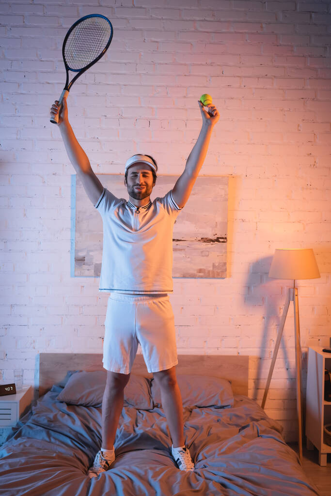 Podekscytowany somnambulista trzyma piłkę tenisową i rakietę na łóżku w nocy  - Zdjęcie, obraz