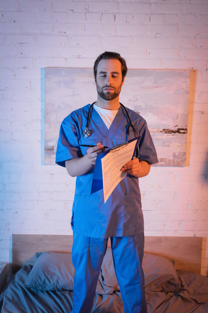 Somnambulist in doctor uniform holding pen and clipboard on bed at night  - Photo, Image