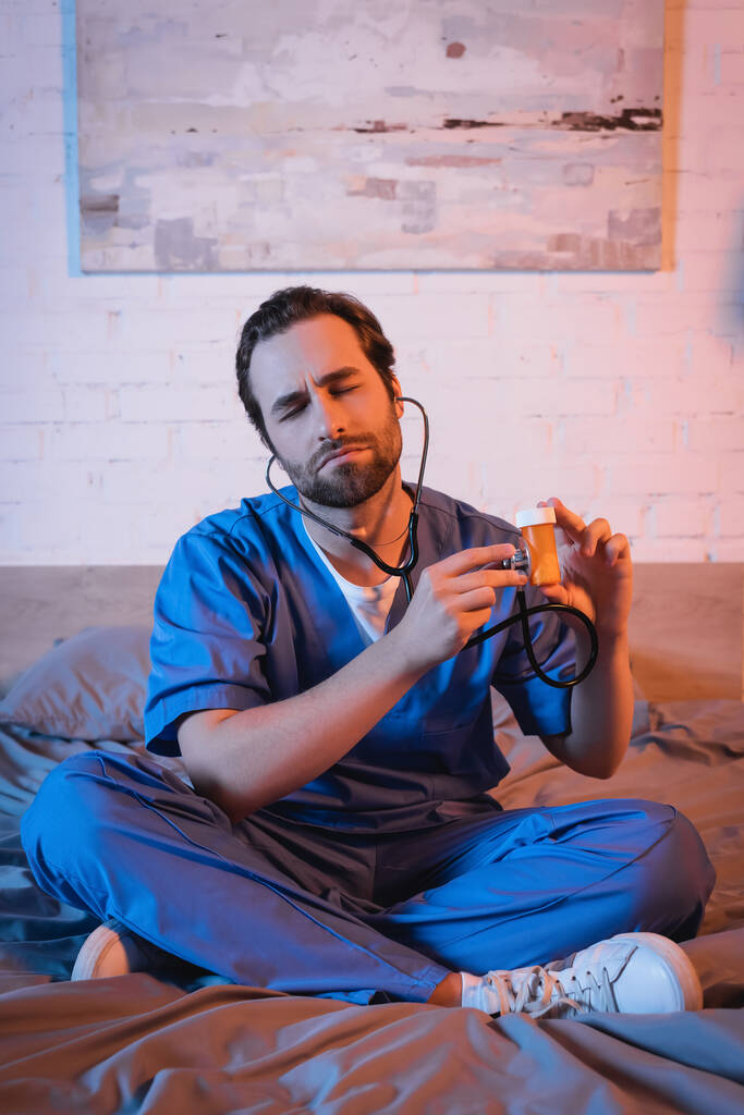 Лунатик в форме врача держит стетоскоп возле таблеток на кровати  - Фото, изображение