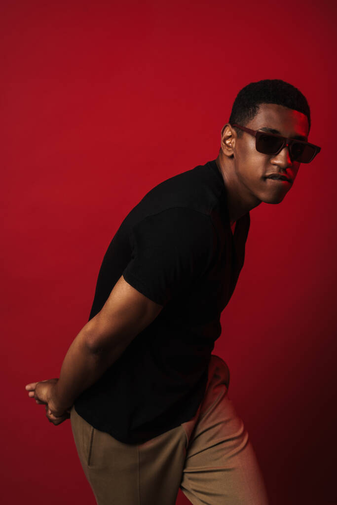 Fiatal fekete férfi napszemüveget visel pózol és a piros fal felett elszigetelt kamerát nézi. - Fotó, kép