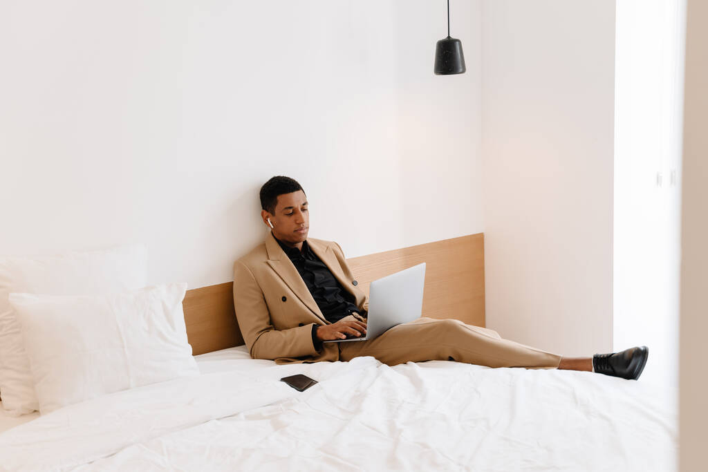 Μαύρος άντρας με ακουστικά που δουλεύει σε λάπτοπ ενώ κάθεται σε χάλια ξενοδοχείο - Φωτογραφία, εικόνα