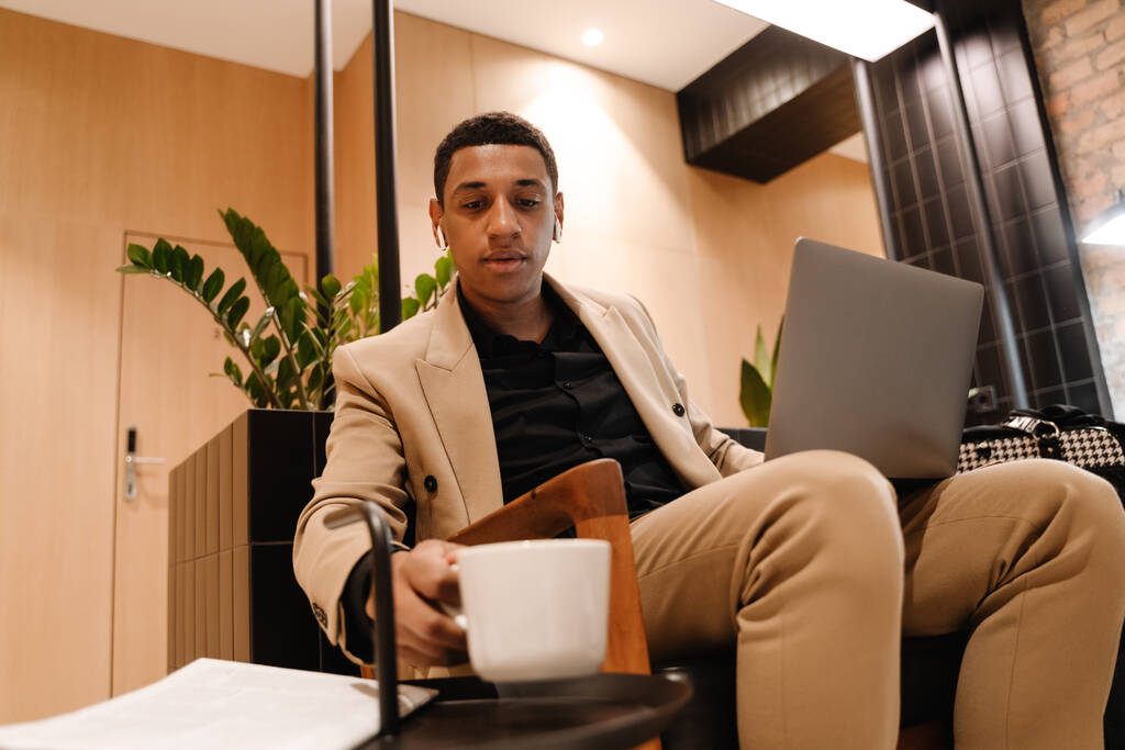 Homme noir dans les écouteurs boire du café tout en travaillant avec un ordinateur portable à l'hôtel - Photo, image