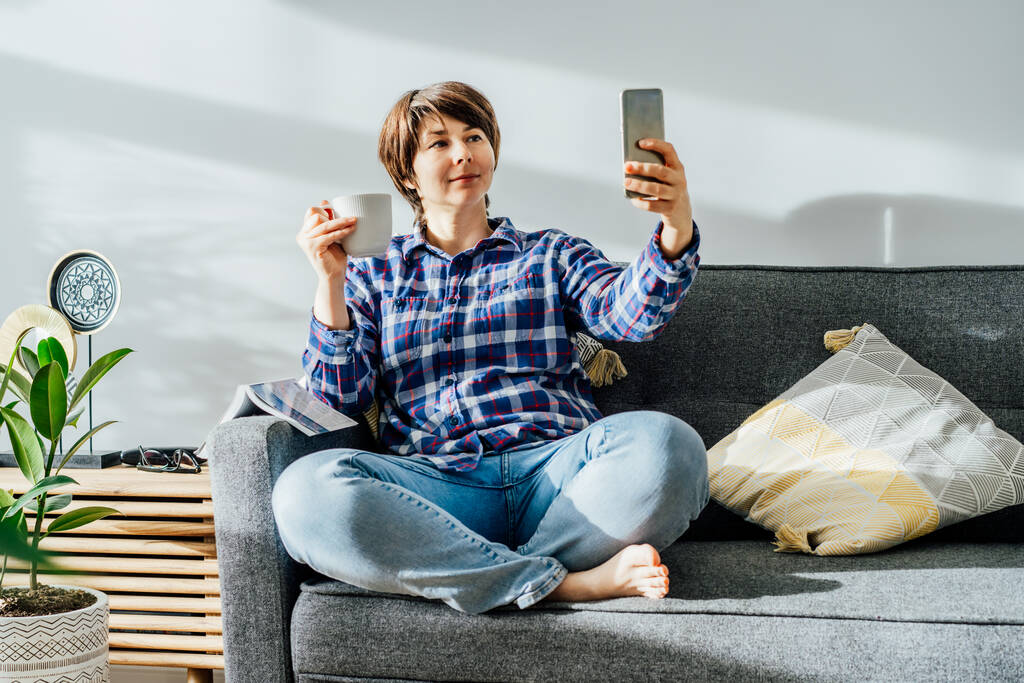 Femme buvant du café ou du thé et faisant selfie par téléphone à la maison assis sur un canapé doux dans le salon intérieur moderne avec des plantes vertes. Influenceur, création de contenu pour les médias sociaux. Espace de copie - Photo, image