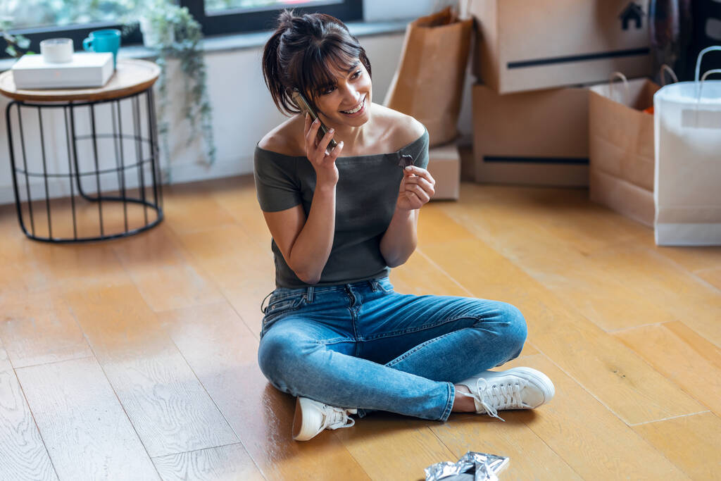 Tiro de joven hermosa mujer con teléfono móvil mientras se come un pedazo de chocolate sentado en el suelo en su nueva casa. - Foto, imagen