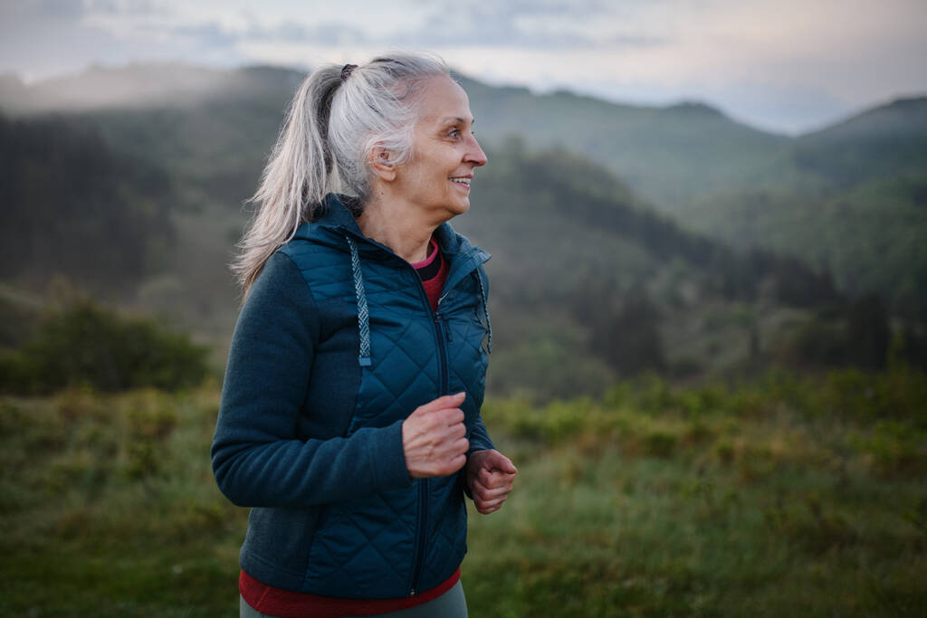 Starší žena jogging v přírodě na časné ráno s mlhou a horami v pozadí. - Fotografie, Obrázek