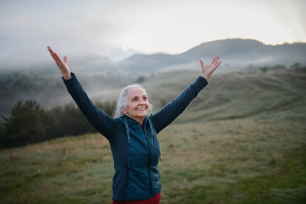 Een oudere vrouw doet ademhalingsoefening in de natuur op de vroege ochtend met mist en bergen op de achtergrond. - Foto, afbeelding