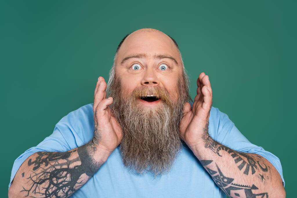 amazed overweight man with beard and tattoos holding hands near face isolated on green - Fotoğraf, Görsel
