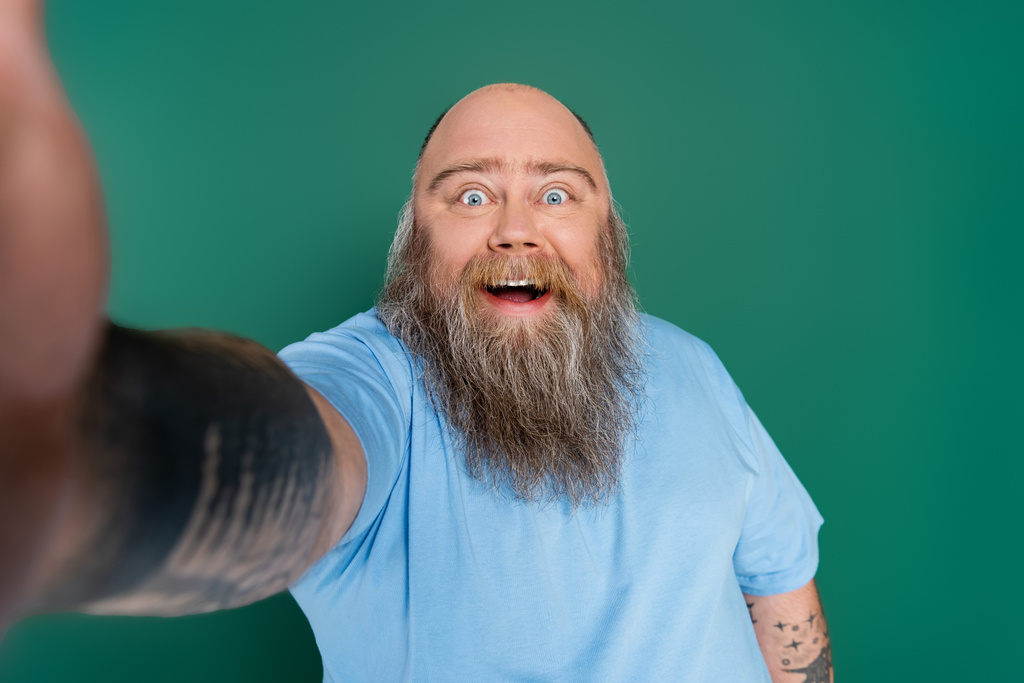 весёлый мужчина с бородой и избыточным весом делает селфи на размытом переднем плане, изолированном на зеленом - Фото, изображение