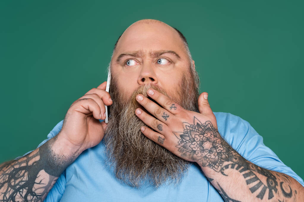 sorprendido hombre tatuado que cubre la boca con la mano mientras habla en el teléfono inteligente aislado en verde - Foto, Imagen