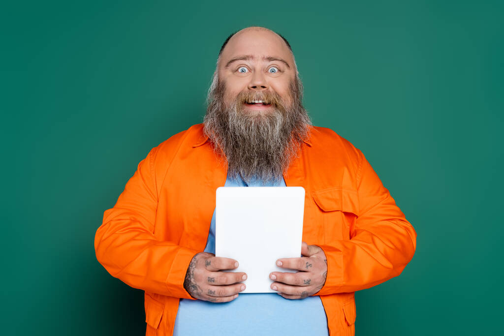 verbaasd en gelukkig plus size man met baard houden digitale tablet geïsoleerd op groen - Foto, afbeelding
