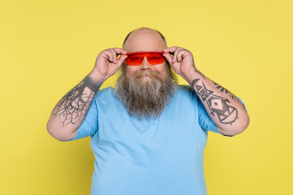 пухлый и татуированный мужчина регулирует модные солнцезащитные очки, улыбаясь в камеру, изолированную на желтом - Фото, изображение