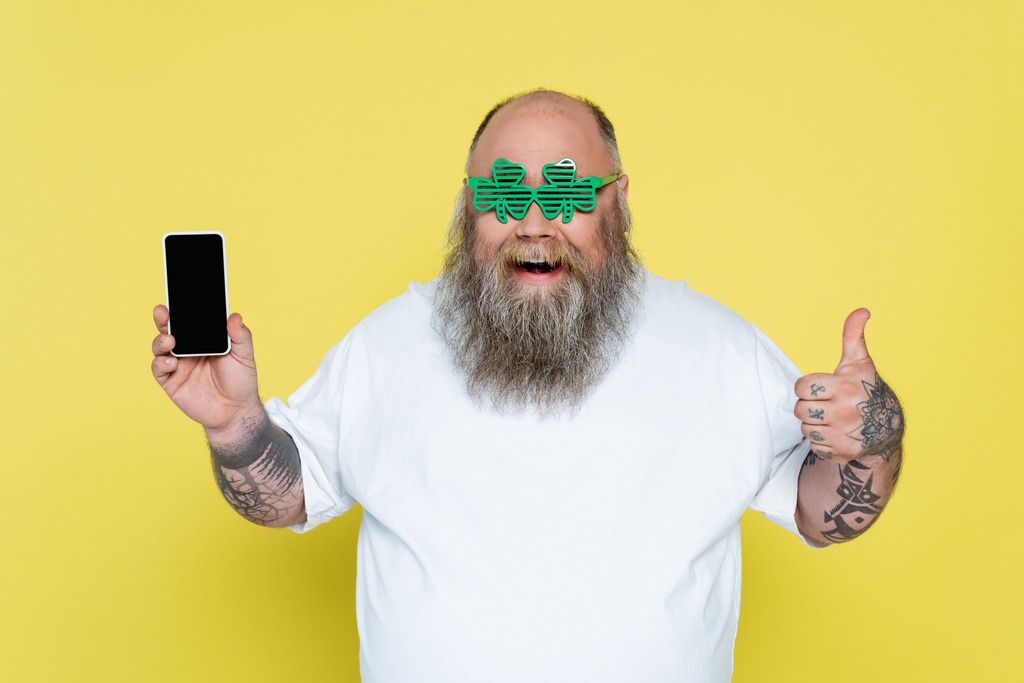 joyful overweight man in clover-shaped glasses showing thumb up while holding smartphone isolated on yellow - Foto, afbeelding