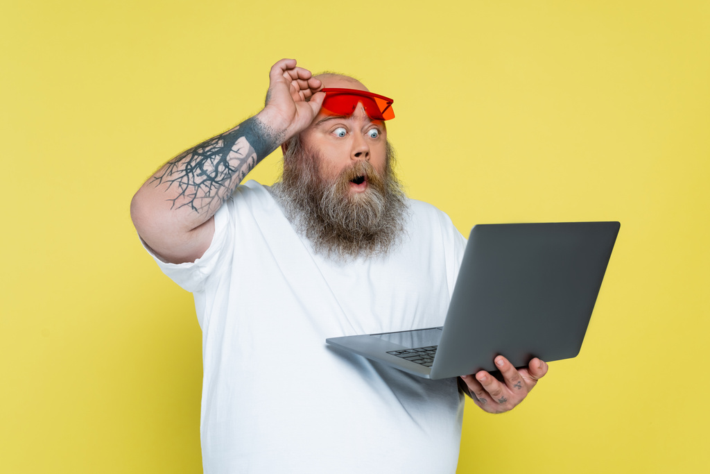 surprised plus size man holding red sunglasses while looking at laptop isolated on yellow - Foto, imagen