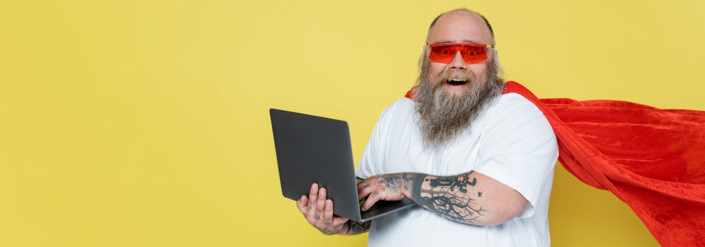wesoły człowiek z nadwagą w czerwonych okularach przeciwsłonecznych i płaszczu superbohatera przy użyciu laptopa izolowanego na żółtym, baner - Zdjęcie, obraz
