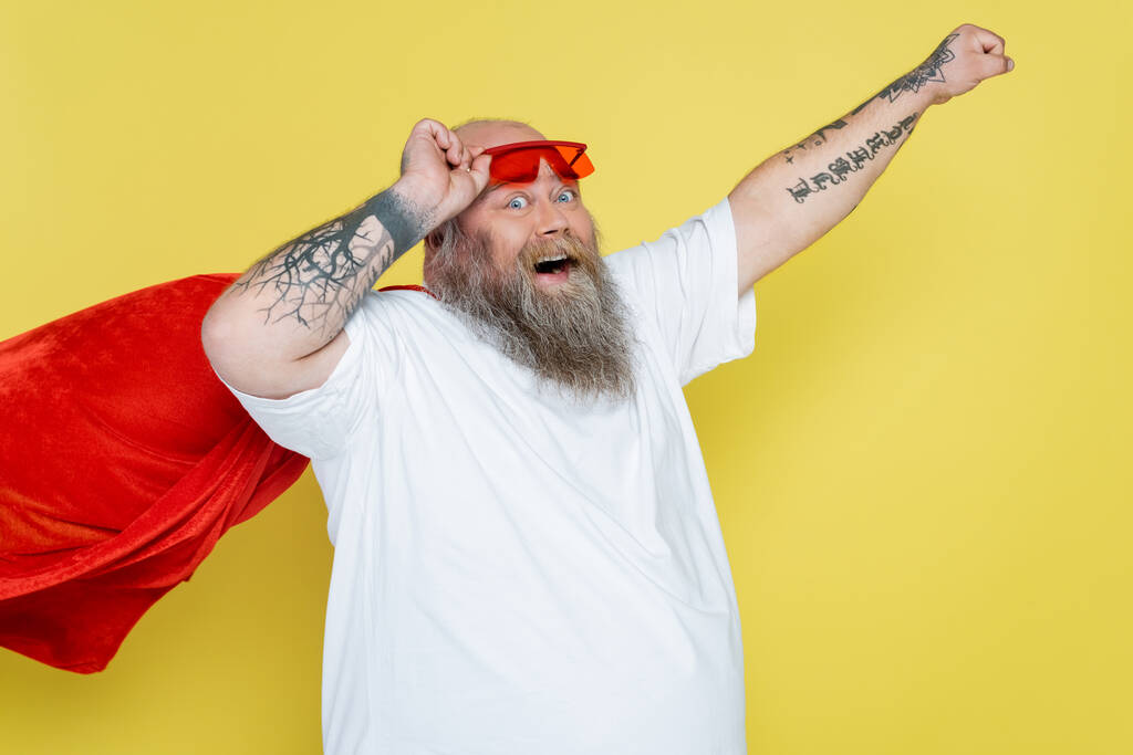 tattooed plus size man in superhero cloak posing with outstretched hand isolated on yellow - Fotó, kép