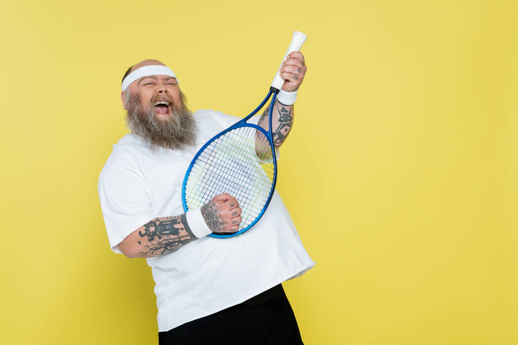 přešťastný plus velikost muž baví s tenisovou raketou izolované na žluté - Fotografie, Obrázek