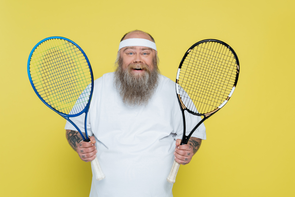 pleased plus size man with beard holding tennis racquets isolated on yellow - Valokuva, kuva