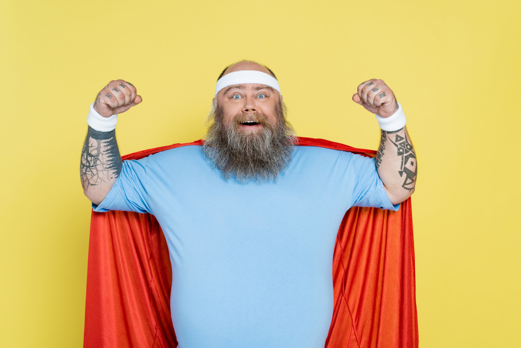 Glatzköpfiger und bärtiger übergewichtiger Sportler im Superheldenmantel, der auf gelb isolierte Macht zeigt - Foto, Bild