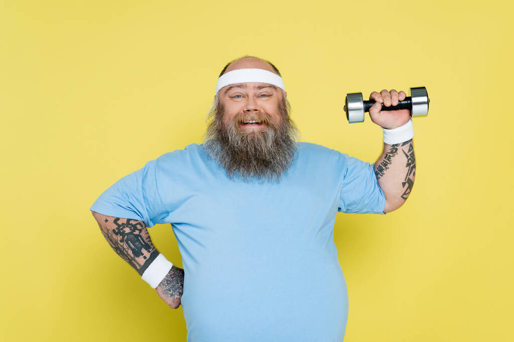 happy overweight man standing with hand on waist and training with dumbbell isolated on yellow - Fotoğraf, Görsel