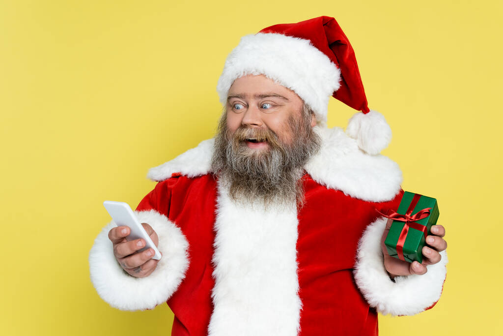 astonished plus size santa claus with present looking at mobile phone isolated on yellow - Foto, immagini