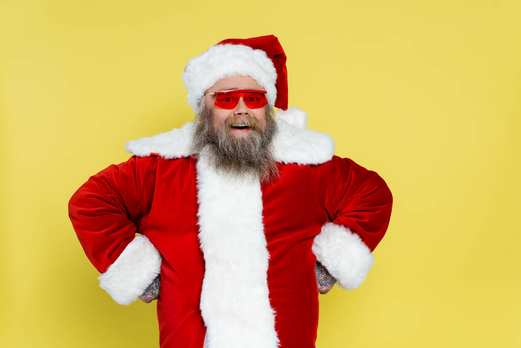 веселый толстый Санта Клаус в красных солнцезащитных очках, стоящих акимбо изолированы на желтый - Фото, изображение