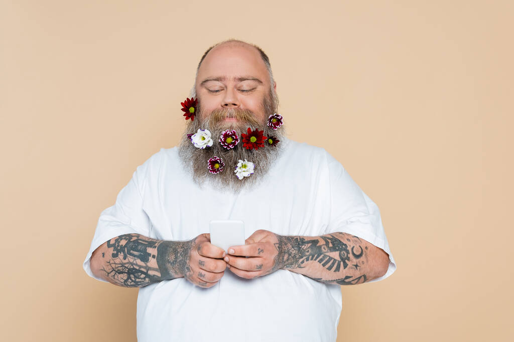 plus velikost muž s květinovou výzdobou na vousy pomocí smartphonu a s úsměvem izolované na béžové - Fotografie, Obrázek