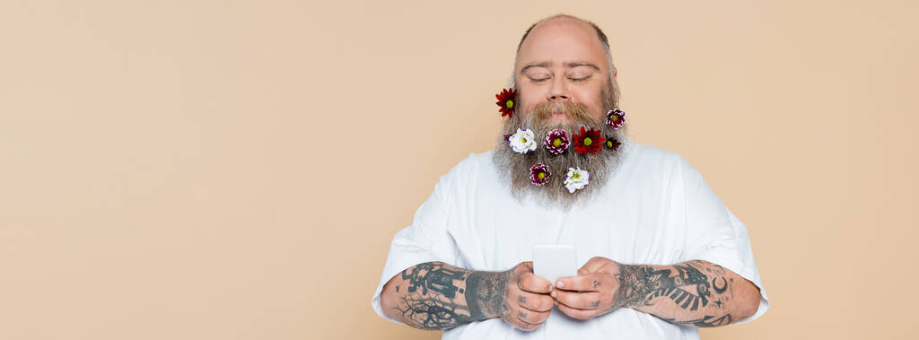 sonriente hombre con sobrepeso con flores en barba usando teléfono celular aislado en beige, pancarta - Foto, imagen