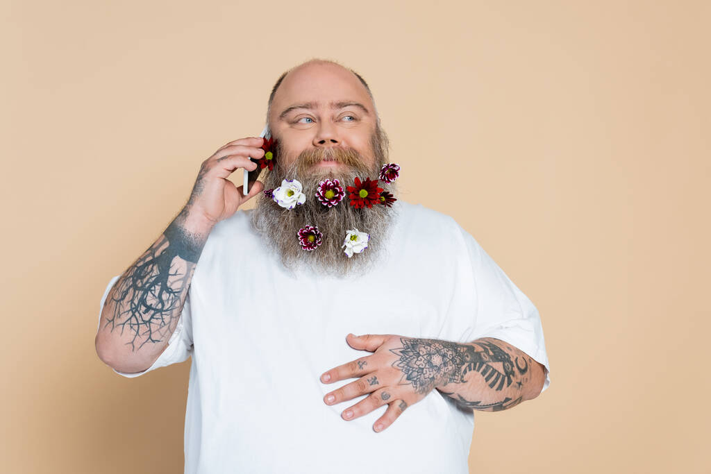 joyful man with flowers in beard talking on mobile phone isolated on beige - 写真・画像
