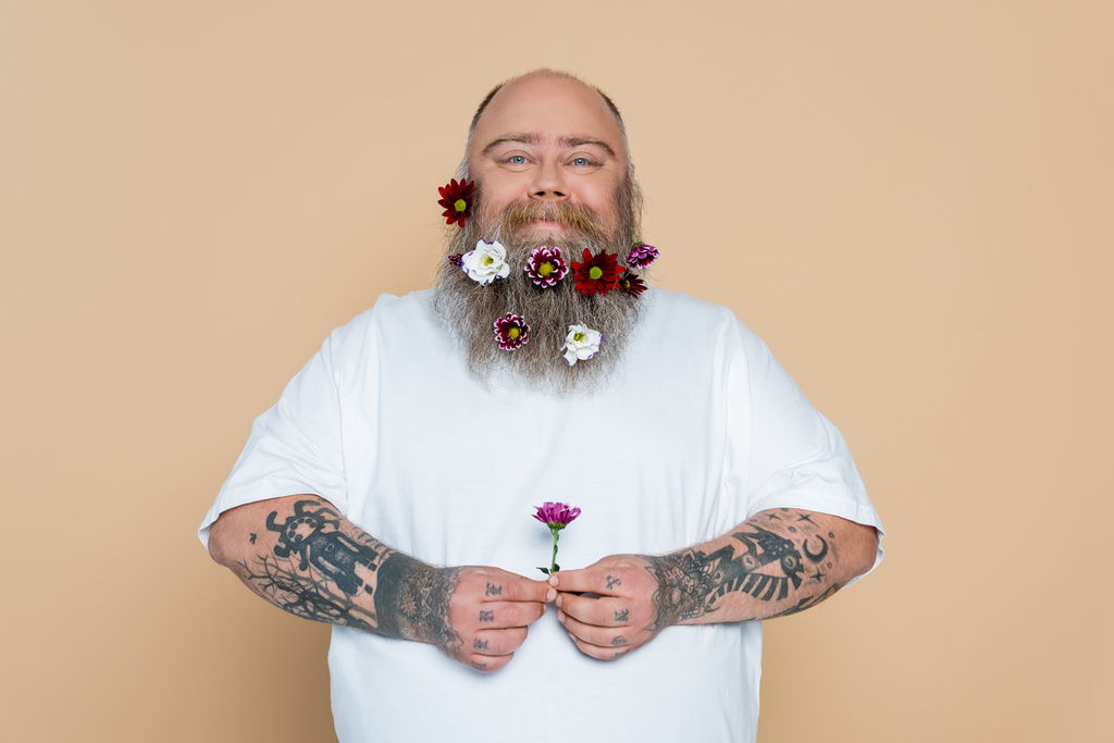 позитивний надмірна вага людини з прикрашеною бородою, що тримає квітку ізольовано на бежевому
 - Фото, зображення