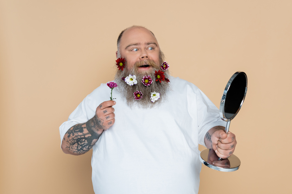 amazed plus size man with flowers in beard looking in mirror isolated on beige - Foto, imagen