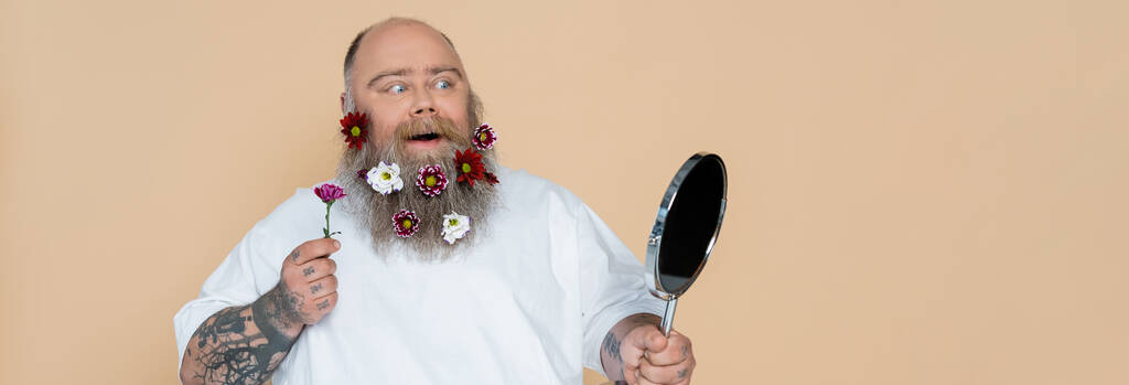 hombre con sobrepeso impresionado con decoración floral en barba mirando en espejo aislado en beige, bandera - Foto, Imagen