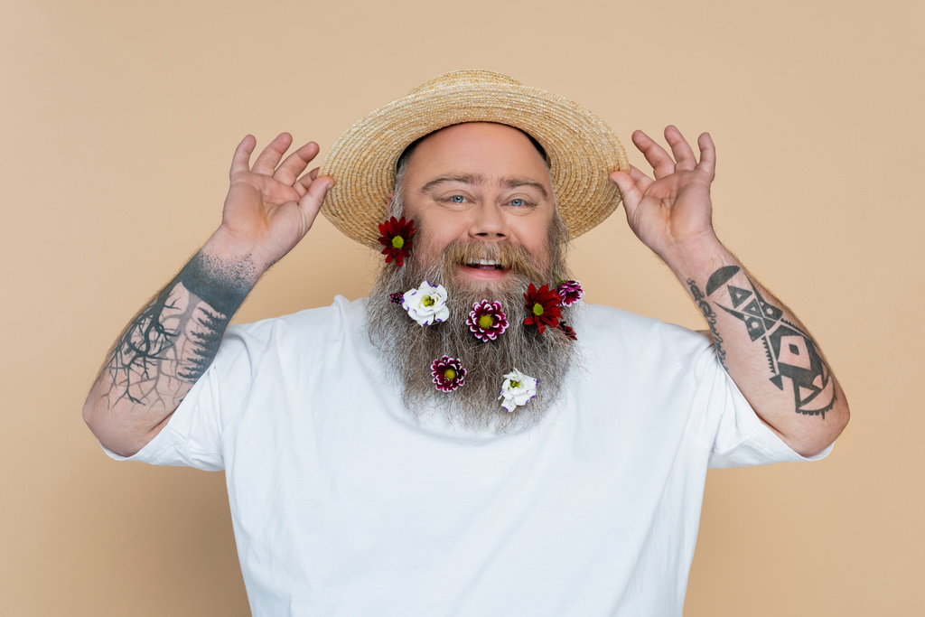 υπέρβαρος άντρας με τατουάζ με διακοσμημένη γενειάδα που προσαρμόζει το ψάθινο καπέλο και χαμογελά στην κάμερα απομονωμένος σε μπεζ - Φωτογραφία, εικόνα