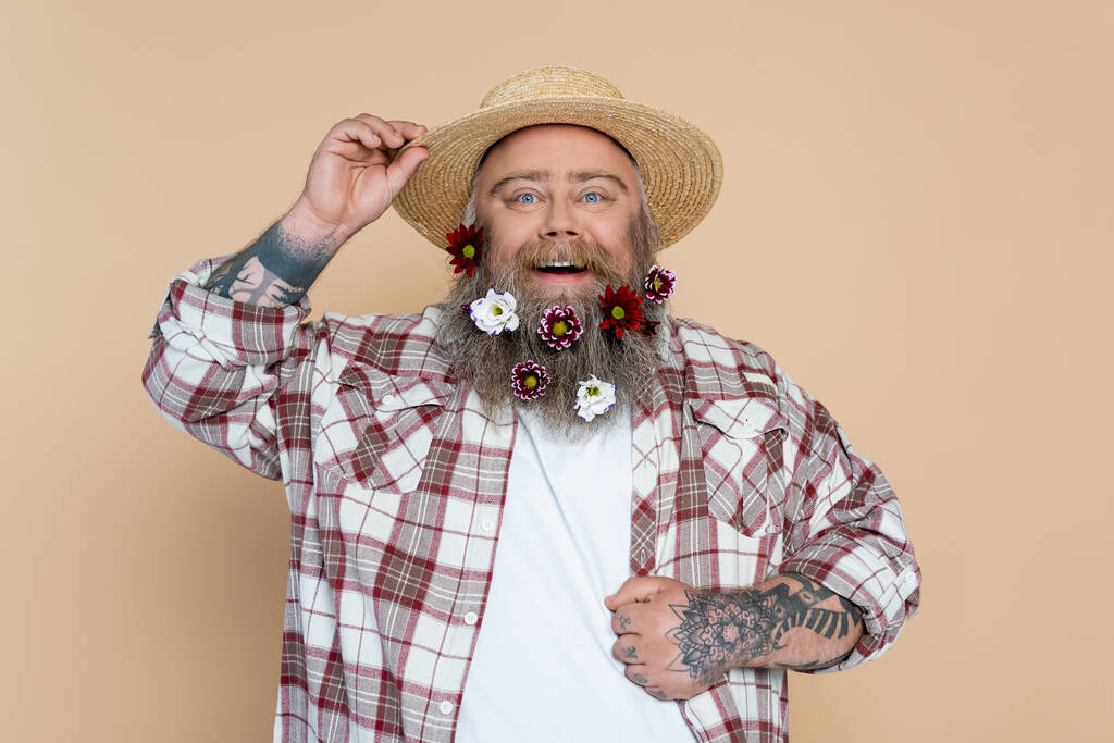 пухлый мужчина с цветами в бороде регулируя соломенную шляпу и улыбаясь в камеру изолированы на бежевый - Фото, изображение