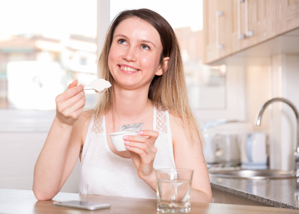 Jonge vrouw zit thuis met yoghurt aan tafel. - Foto, afbeelding