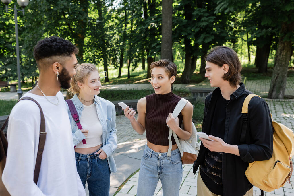 Estudiante alegre con smartphone hablando con amigos interracial en el parque  - Foto, imagen
