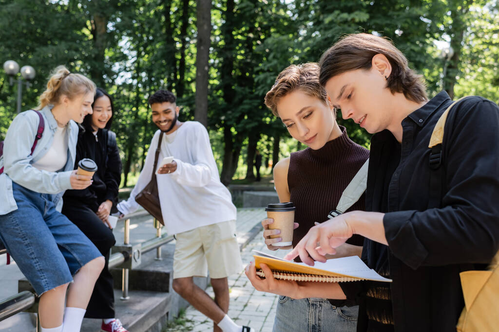 Estudiantes mirando cuaderno cerca borrosa amigos multiétnicos con café y teléfonos inteligentes en el parque  - Foto, imagen