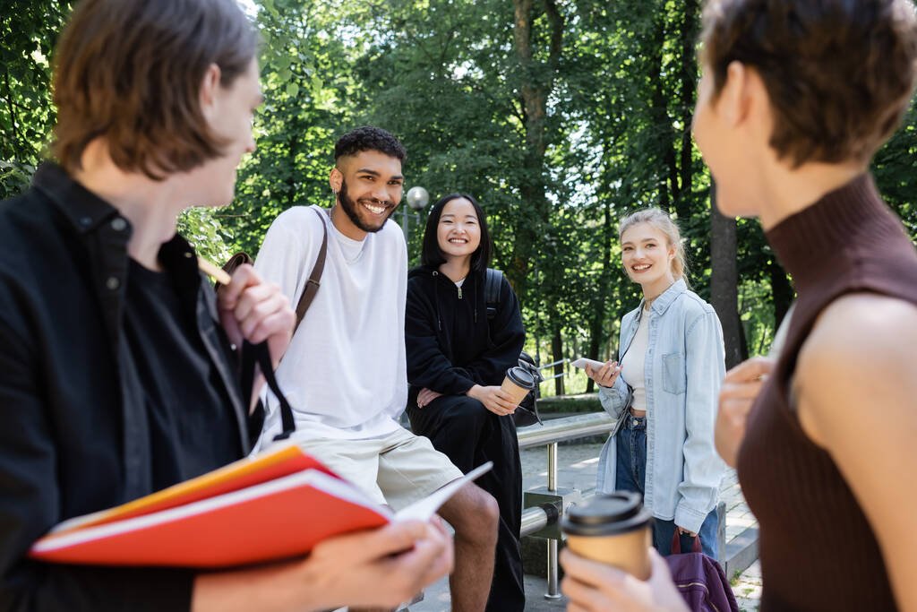Cheerful multicultural students with smartphone and takeaway drink looking at blurred friends in park  - 写真・画像
