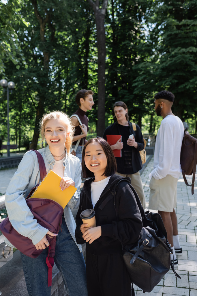 Межрасовые студенты с ноутбуком и кофе, чтобы пойти улыбаясь в камеру рядом размытые друзья в парке  - Фото, изображение