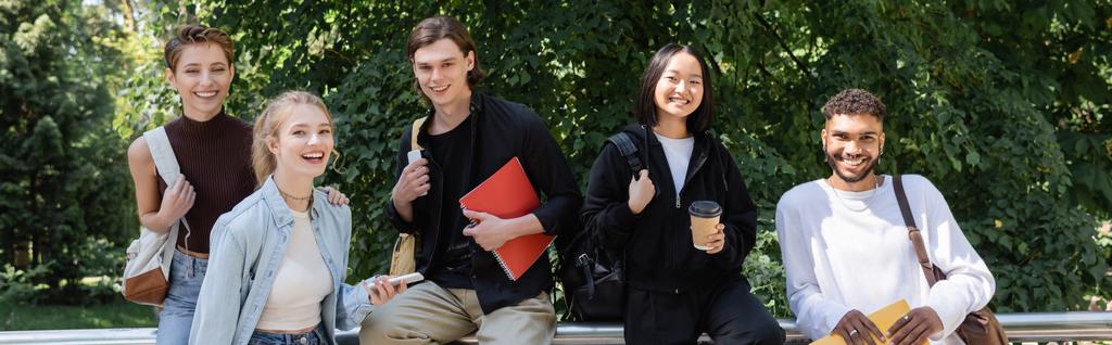 Cheerful interracial students with backpacks and notebooks looking at camera in park, banner  - Photo, Image