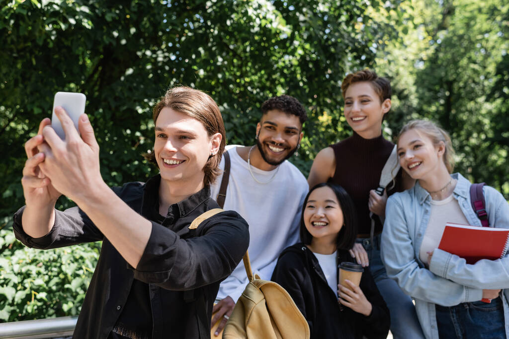 Studente sorridente scattare selfie su smartphone vicino amici interrazziale nel parco  - Foto, immagini