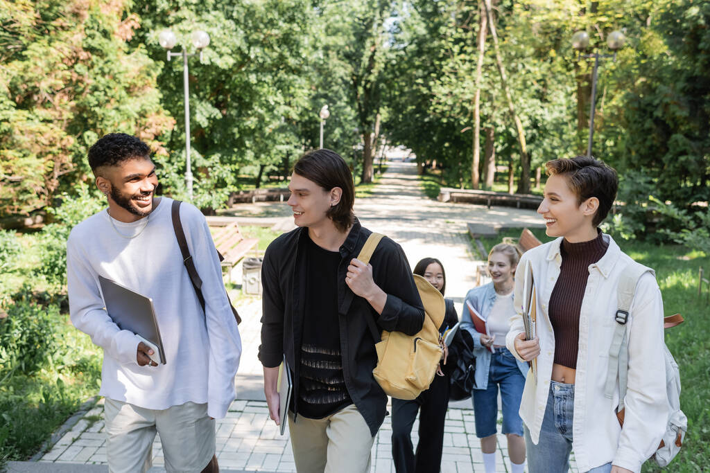 Étudiants interraciaux avec appareils et sacs à dos marchant dans le parc  - Photo, image