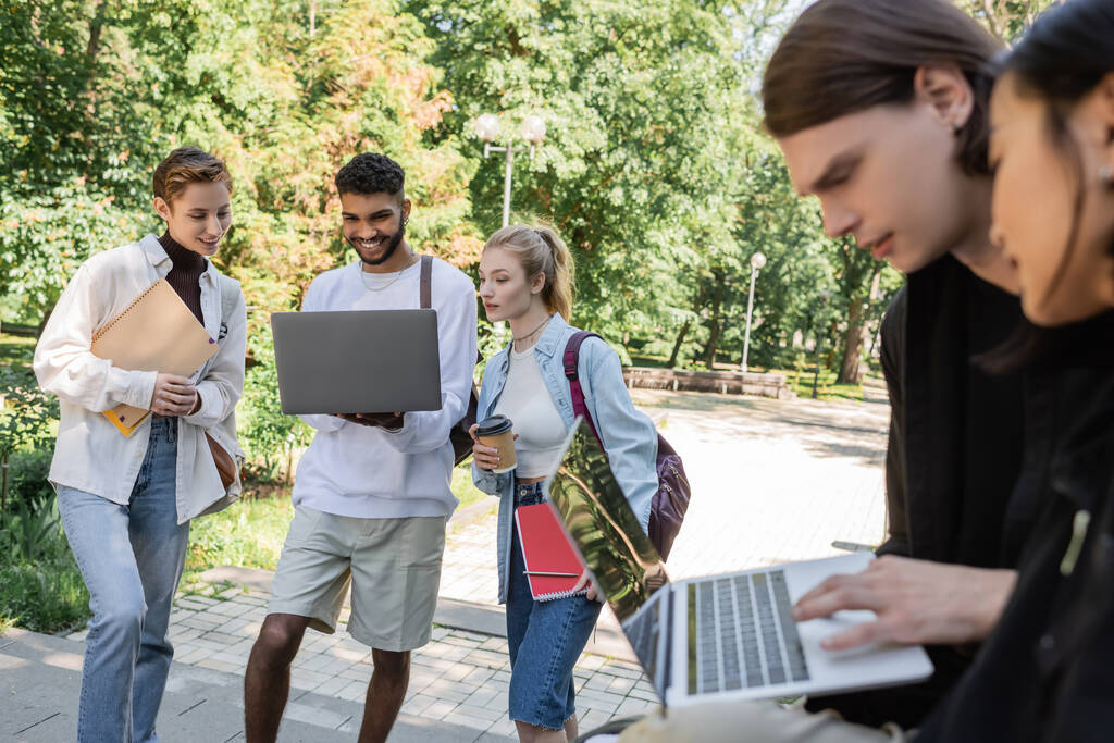 African american student holding laptop near friends with notebooks in park  - 写真・画像