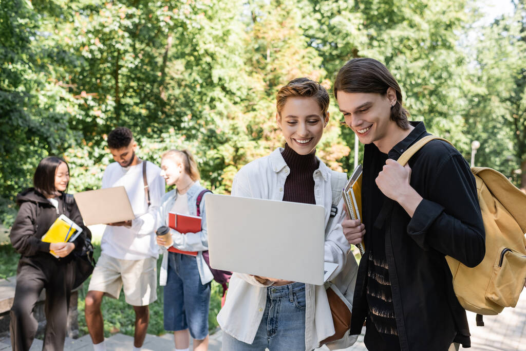 Cheerful students using laptop near blurred multicultural friends in park  - Foto, Bild