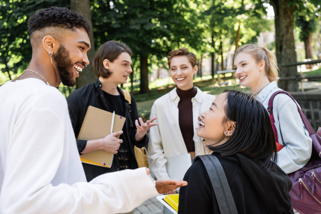 Усміхнені міжрасові студенти розмовляють біля розмитих друзів з рюкзаками в літньому парку
  - Фото, зображення