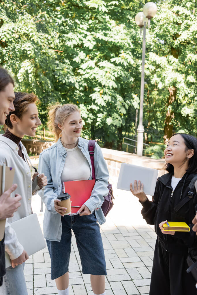 Uśmiechnięci studenci z notatnikami patrzący na azjatyckiego przyjaciela z cyfrowym tabletem w parku  - Zdjęcie, obraz